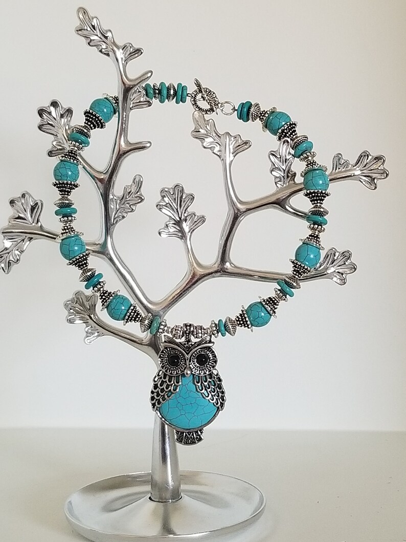 May include: A turquoise and silver owl necklace with turquoise beads. The necklace is displayed on a silver tree-shaped stand.