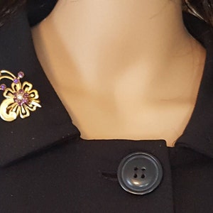 Puede incluir: Un broche dorado con un dise&ntilde;o floral y piedras de imitaci&oacute;n de color p&uacute;rpura. El broche est&aacute; prendido a un cuello negro.