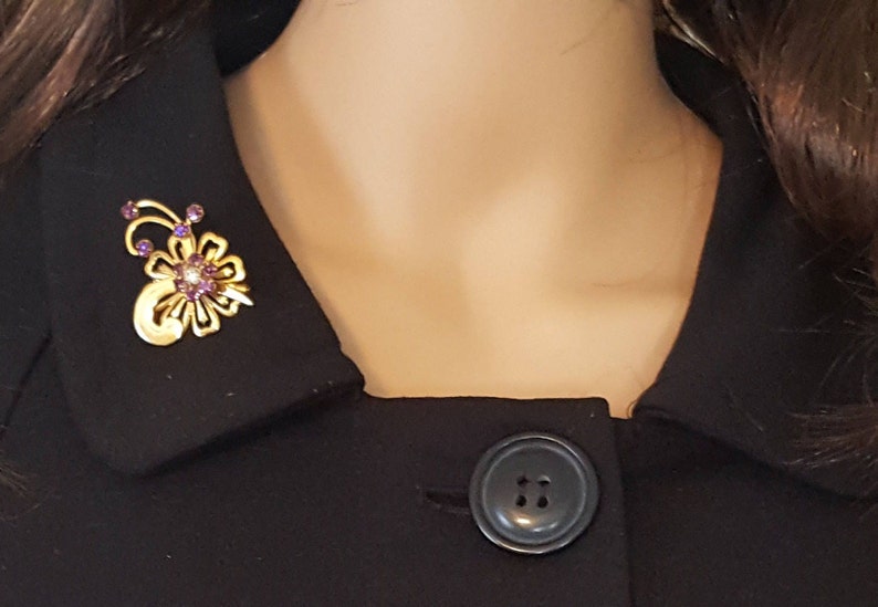 Puede incluir: Un broche dorado con un dise&ntilde;o floral y piedras de imitaci&oacute;n de color p&uacute;rpura. El broche est&aacute; prendido a un cuello negro.