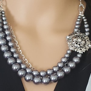 May include: A three-strand gray pearl necklace with a silver-tone filigree pendant. The necklace features a chain detail and a decorative clasp. The pearls vary in size, creating a layered effect. A classic piece of jewelry.