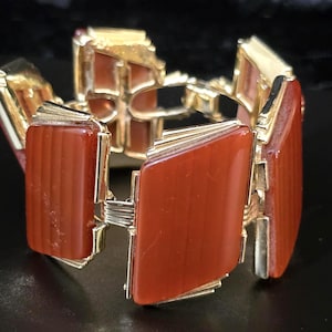 May include: A vintage bracelet featuring rectangular, reddish-brown stones set in a gold-tone metal frame. The bracelet has a hinged design with multiple segments, creating a bold and eye-catching accessory. The stones have a ribbed texture.