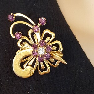 Puede incluir: Un broche dorado con dise&ntilde;o de flor. La flor tiene un centro de piedras de cristal transparentes y p&eacute;talos hechos de piedras de cristal moradas. El broche tiene un tallo curvo con una piedra de cristal morada en la parte superior.
