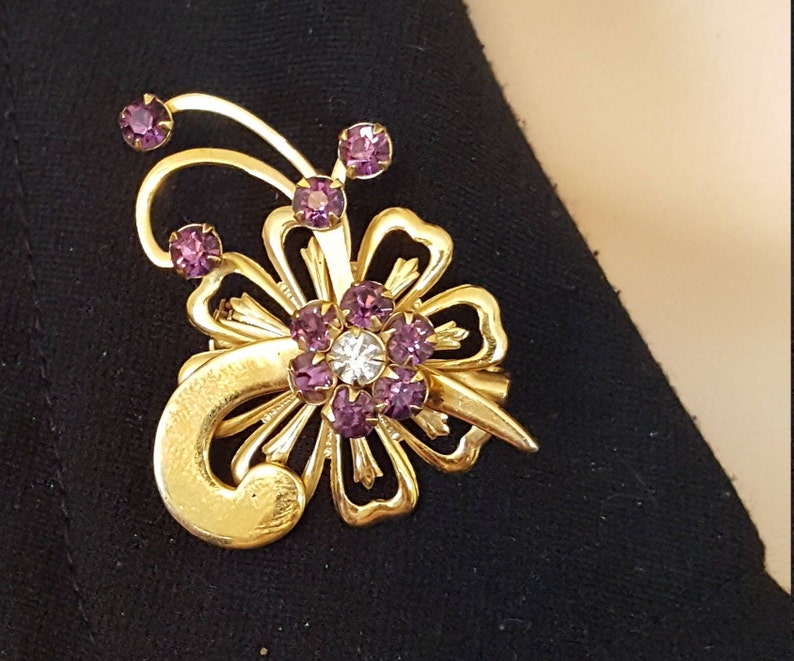 Puede incluir: Un broche dorado con dise&ntilde;o de flor. La flor tiene un centro de piedras de cristal transparentes y p&eacute;talos hechos de piedras de cristal moradas. El broche tiene un tallo curvo con una piedra de cristal morada en la parte superior.