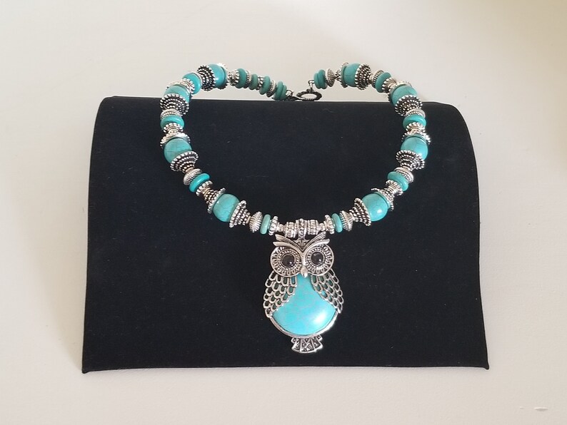 May include: A turquoise and silver necklace with an owl pendant. The owl has black eyes and is surrounded by turquoise stones.