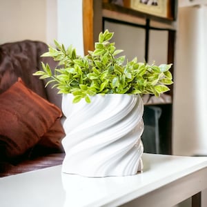 Può includere: Un vaso di ceramica bianca con un design a spirale, che contiene una pianta verde a foglie. Il vaso è appoggiato su un tavolo bianco.