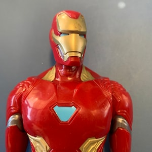 May include: A red and gold action figure of Iron Man, a superhero from the Marvel universe. The figure has a blue light in its eyes and a blue arc reactor on its chest.