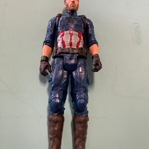 May include: A plastic action figure of Captain America wearing a blue and red costume with a brown belt and brown boots. The figure has a beard and short brown hair.