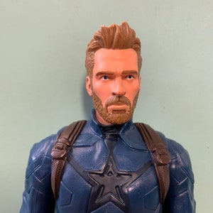 May include: A plastic action figure of Captain America wearing a blue and red costume with a star on the chest. He has a brown beard and brown hair.