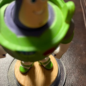 May include: A green and purple toy figure of Buzz Lightyear from the Toy Story franchise. The figure is standing on a brown wooden base with a clear plastic ring around it.