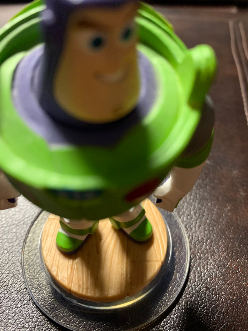 May include: A green and purple toy figure of Buzz Lightyear from the Toy Story franchise. The figure is standing on a brown wooden base with a clear plastic ring around it.