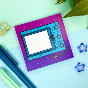 May include: A square sticker featuring a blue and purple design reminiscent of a retro handheld device. The sticker has a white screen and a chain detail. A gold paper clip, blue pen, and star-shaped beads are also visible.
