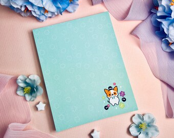 May include: A light blue notebook with a white pattern of stars and a cartoon illustration of a dog holding a blue ball. The dog is surrounded by small purple and pink flowers.