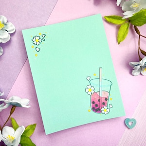Könnte beinhalten: Eine hellblaue Karte mit einer Illustration von rosa und weißem Bubble Tea und weißen Blumen in der Ecke. Die Karte hat einen hellgrünen Hintergrund.