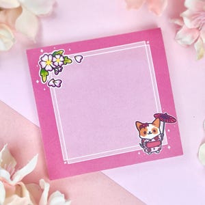 May include: Pink sticky note with a white border and a floral design in the top corner. The note has a white frame with a cartoon image of a dog wearing a kimono and holding an umbrella.