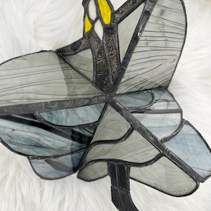 May include: Stained glass dragonfly suncatcher with gray and blue wings, yellow and white accents, and a black frame. The dragonfly is suspended by a silver chain. The wings have a striped design.