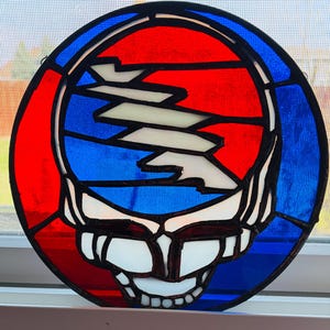 May include: Round stained glass art piece featuring a stylized skull design. The artwork uses red, blue, white, and black colors. The design is reminiscent of a popular band's logo.