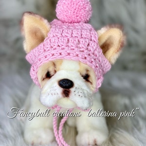 May include: A pink crocheted hat with a pom-pom on top. The hat is on a white stuffed dog with brown ears. The text "fancybull creations "ballerina pink" is written below the dog.