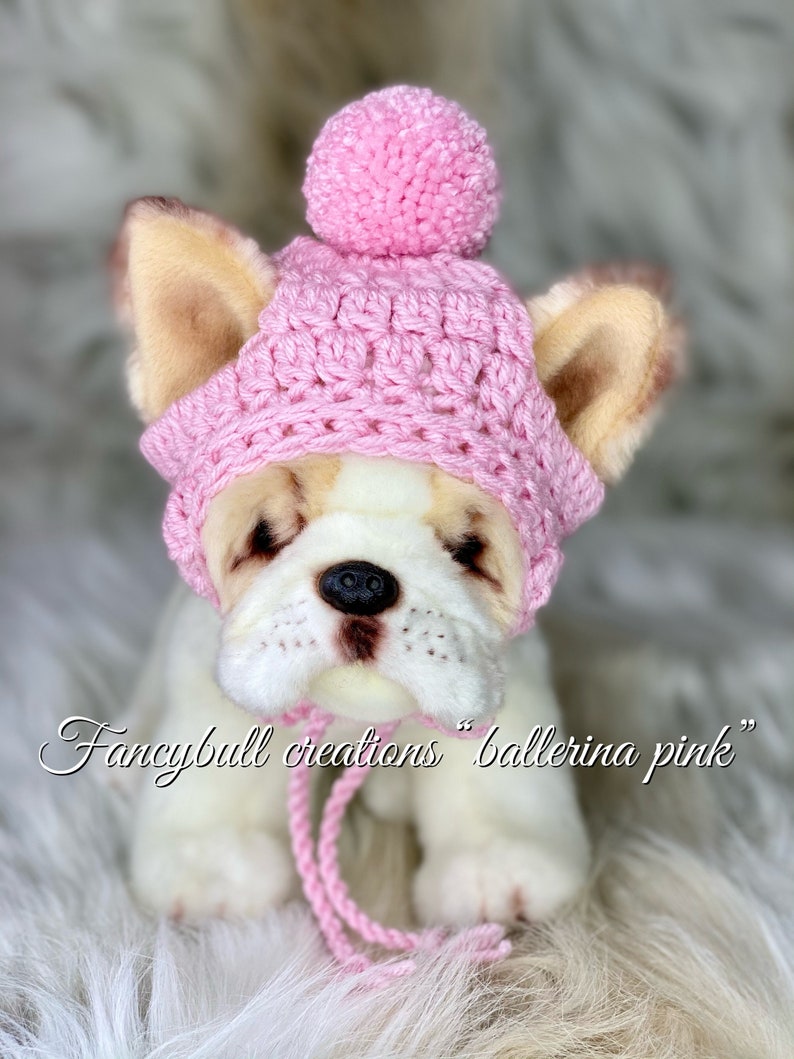 May include: A pink crocheted hat with a pom-pom on top. The hat is on a white stuffed dog with brown ears. The text "fancybull creations "ballerina pink" is written below the dog.