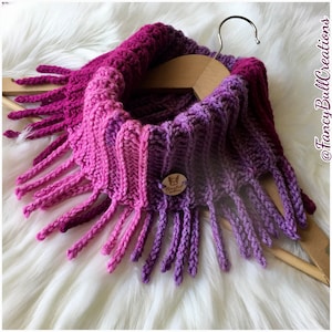 Może przedstawiać: Ombreowy szalik typu infinity w kolorze fioletowym i różowym z frędzlami. Szalik jest prezentowany na drewnianym wieszaku. Etykieta na szaliku głosi "Fancy Bull Creations".