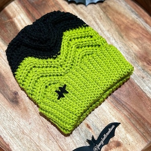 May include: A green and black crocheted hat with a star design. The hat is on a wooden surface with a black bat cutout.