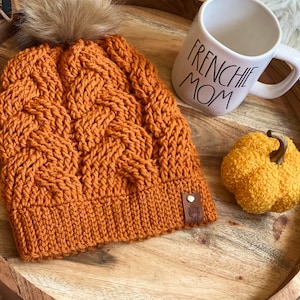 Könnte beinhalten: Eine braune Strickmütze mit einem Kunstfellbommel oben. Die Mütze hat ein Zopfmuster und ein braunes Leder-Etikett mit einem kleinen Bild eines Hundes. Die Mütze liegt auf einem Holztablett mit einer weißen Tasse, auf der "Frenchie Mom" steht, und einem gelben Deko-Kürbis.