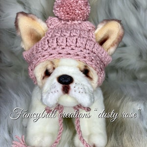 May include: A pink crocheted dog hat with a pom pom on top. The hat is on a white stuffed dog with brown ears. The text "fancyball creations "dusty rose" is below the dog.