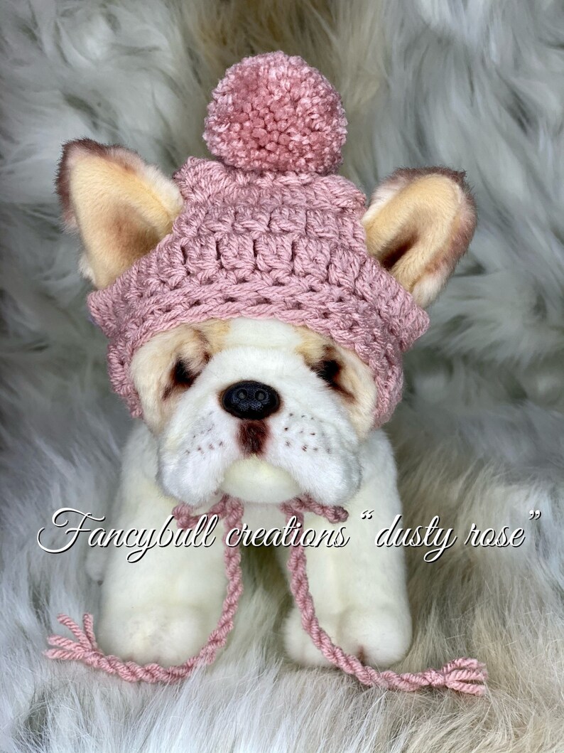 May include: A pink crocheted dog hat with a pom pom on top. The hat is on a white stuffed dog with brown ears. The text "fancyball creations "dusty rose" is below the dog.