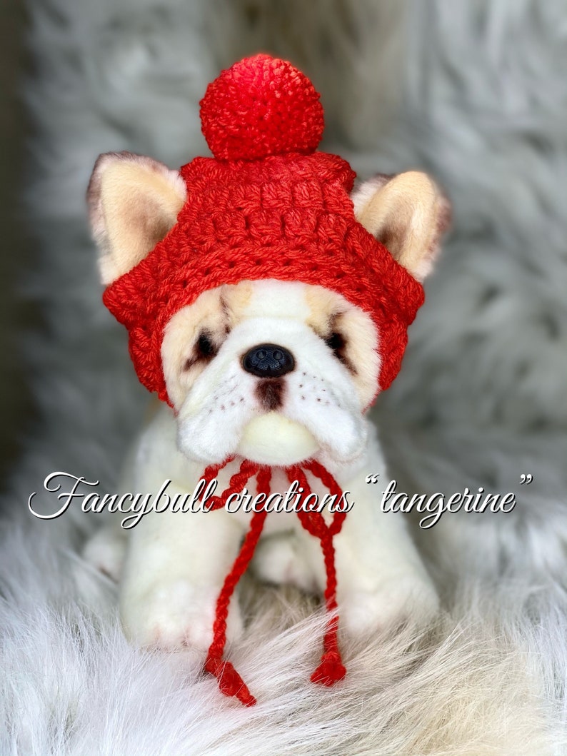 May include: A white stuffed dog wearing a red crocheted hat with a pom-pom on top. The hat has a red string tie. The dog is sitting on a white fluffy surface. "Fancybull creations "tangering""
