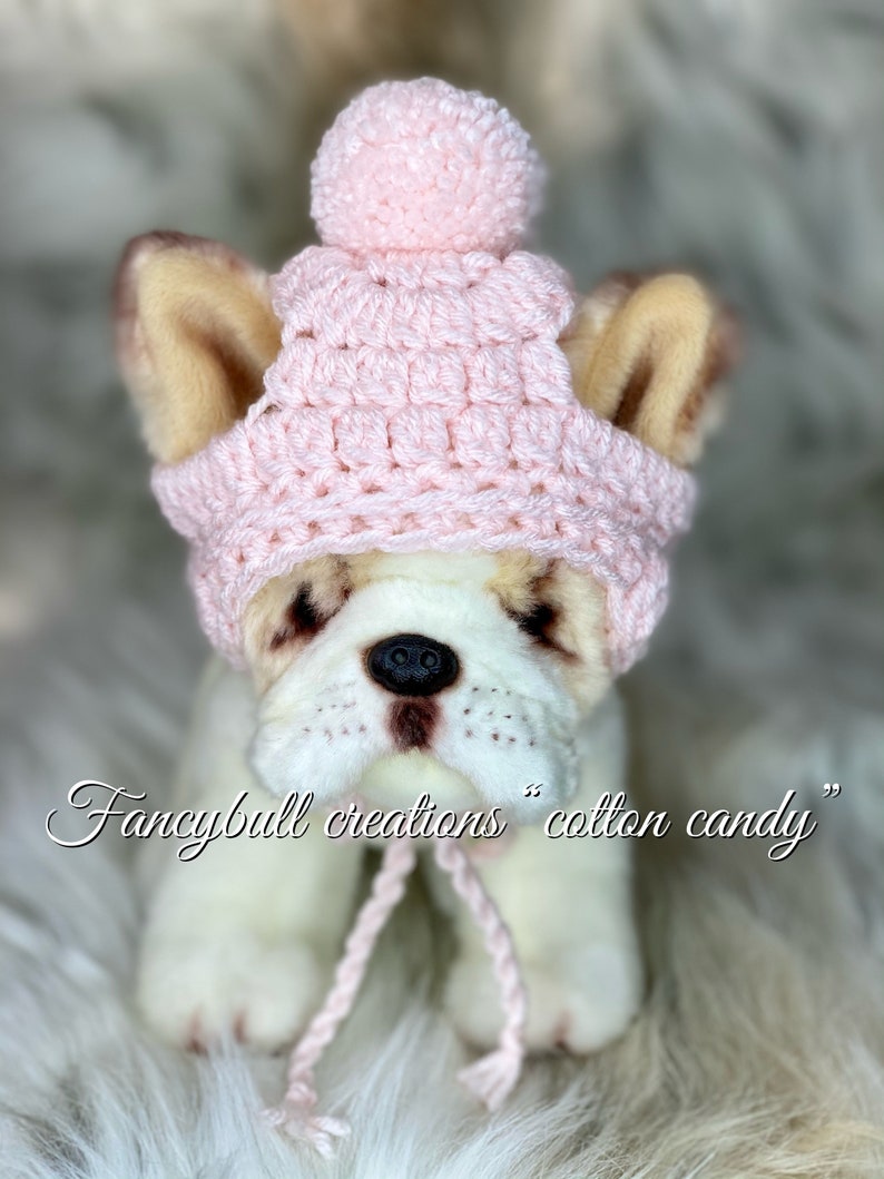 May include: A pink crocheted hat with a pom-pom on top. The hat is designed for a small dog and has a long, dangling tie. The hat is sitting on a white furry surface. "Fancybull creations cotton candy" is written in cursive below the hat.