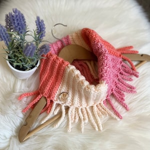 May include: A pink, white, and coral striped crocheted infinity scarf with fringe. The scarf is displayed on a wooden hanger and has a small wooden tag with the text "Simply Crafted" on it.