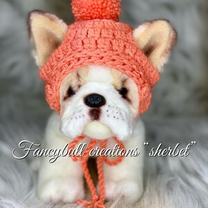 May include: A white stuffed dog wearing a crocheted coral pink hat with a pom-pom on top. The hat has a matching coral pink scarf tied around the dog's neck. The dog is sitting on a white fluffy surface. The text "fancybull creations "sherbet" is written below the dog.