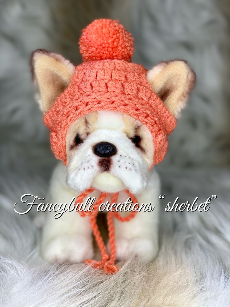 May include: A white stuffed dog wearing a crocheted coral pink hat with a pom-pom on top. The hat has a matching coral pink scarf tied around the dog's neck. The dog is sitting on a white fluffy surface. The text "fancybull creations "sherbet" is written below the dog.