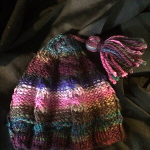 May include: A hand-knitted hat in a mix of colors including purple, pink, blue, and green. The hat has a textured pattern and a tassel in matching colors. The hat is made of soft yarn and is suitable for cold weather.