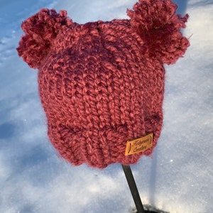 May include: A burgundy knitted hat with two fluffy pom-poms on top. The hat has a brown leather label that says "Haleymoon Creations".