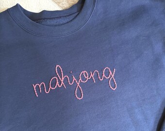 May include: Navy blue sweatshirt with the word "mahjong" embroidered in pink cursive script. The crew neck sweatshirt is made of a soft material.