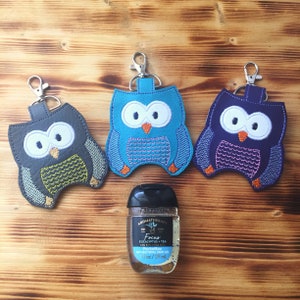 May include: Three owl-shaped keychains in gray, blue, and purple, each with a silver clasp. The keychains are embroidered with white eyes and orange beaks. A bottle of PocketBac anti-bacterial hand gel is in front of the keychains.
