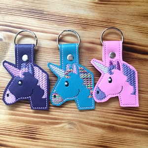 May include: Three unicorn keychains in purple, teal, and pink. Each keychain features a stitched unicorn design with a silver metal keyring and a matching leather strap. The unicorns have embroidered manes and smiling faces.