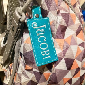 May include: A teal leather keychain tag with the name "Jacobi" embroidered in white. The tag is attached to a gray backpack with a geometric pattern in shades of purple, peach, and white. The keychain has a silver metal ring.