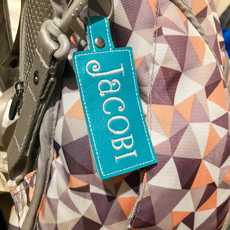 May include: A teal leather keychain tag with the name "Jacobi" embroidered in white. The tag is attached to a gray backpack with a geometric pattern in shades of purple, peach, and white. The keychain has a silver metal ring.