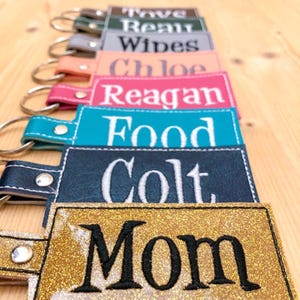 May include: A collection of personalized keychains in various colors, including gold glitter, teal, pink, and navy. Each keychain features a name or word embroidered in white, such as "Mom," "Colt," and "Food." The keychains are attached to silver key rings.
