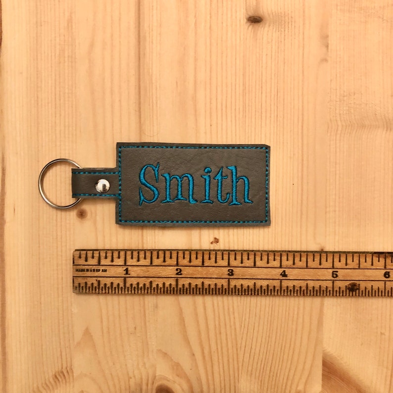 May include: A rectangular gray leather keychain with the name "Smith" embroidered in blue. The keychain has a blue stitched border and a silver key ring attached. A wooden ruler is placed next to the keychain.