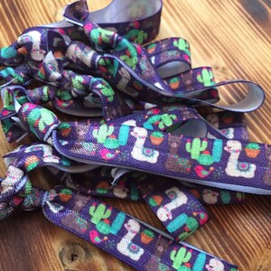 Puede incluir: Un conjunto de coleteros morados con un estampado repetido de llamas y cactus en verde, marrón y rosa. Los coleteros están anudados y colocados sobre una superficie de madera.
