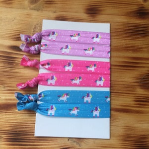 Puede incluir: Un conjunto de cinco gomas para el pelo coloridas con un estampado de unicornios. Las gomas son de tonos rosas y azules, con unicornios blancos y crines arcoíris. Presentadas sobre una tarjeta blanca.