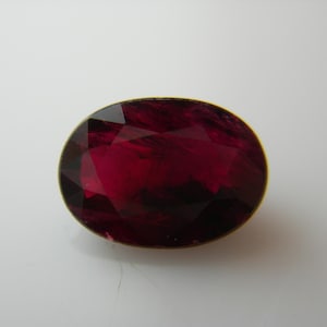 May include: An oval-shaped, faceted, deep red gemstone with a gold-colored bezel. The gemstone has a brilliant cut, reflecting light and creating a sparkling effect. The rich color and cut suggest it could be a ruby or garnet, suitable for jewelry.