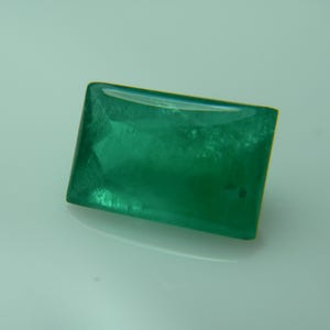 May include: A rectangular, emerald-green gemstone with a polished, reflective surface. The gem has a slightly rounded shape and a subtle internal texture, suggesting it may be a natural stone. The color is a rich, vibrant green.