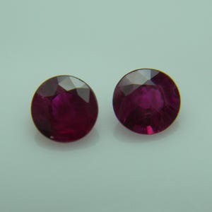 Puede incluir: Dos piedras preciosas redondas y facetadas de un color rojo magenta intenso. Cada piedra presenta un corte circular con múltiples facetas triangulares que reflejan la luz, creando una apariencia brillante y centelleante en la superficie.