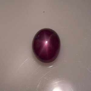 May include: A polished, oval-shaped, deep burgundy gemstone with a six-ray star effect. The gem has a smooth, reflective surface and a prominent star pattern emanating from the center. The stone is set against a neutral background.