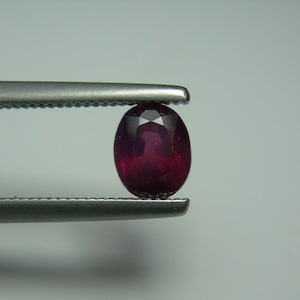 Puede incluir: Una gema de rubí ovalada y facetada, sujeta con unas pinzas plateadas. El rubí es de color rojo intenso con una superficie pulida que refleja la luz. Las pinzas se encuentran sobre un fondo gris claro.