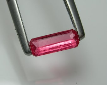 May include: A faceted, rectangular, pink gemstone is held by a pair of silver tweezers. The gemstone has a vibrant, translucent color and a rectangular shape with cut edges. The tweezers are in sharp focus.