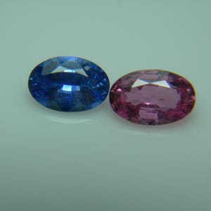 Peut inclure: Deux pierres précieuses de forme ovale. L'une est d'un bleu profond, l'autre d'un rose vif. Les deux pierres ont une surface polie et facettée qui reflète la lumière. Les pierres sont posées sur un fond clair.